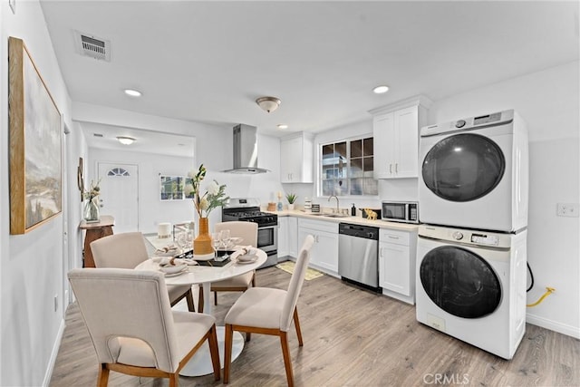 kitchen with wall chimney range hood, sink, white cabinetry, stacked washing maching and dryer, and appliances with stainless steel finishes