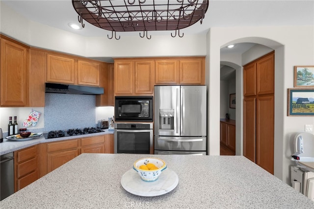 kitchen with arched walkways, light stone counters, under cabinet range hood, black appliances, and brown cabinetry