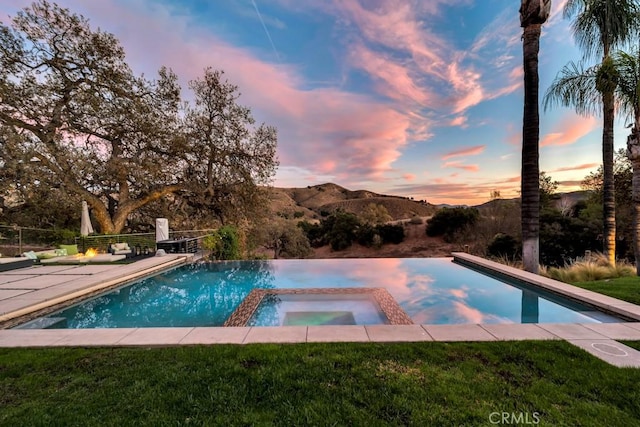 pool at dusk featuring a lawn, a patio area, and an in ground hot tub