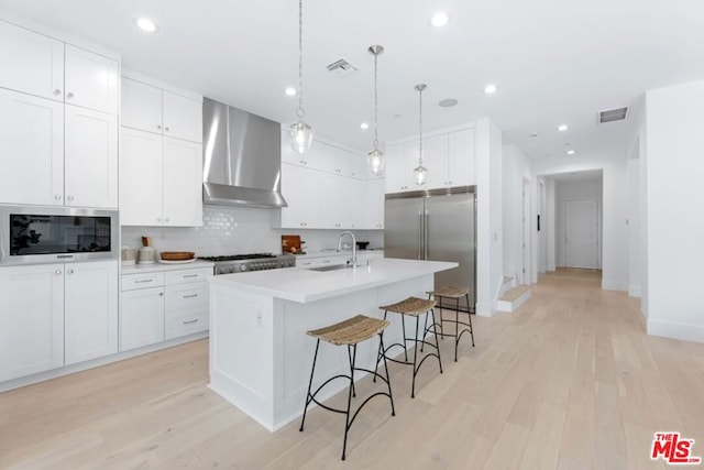 kitchen with white cabinetry, wall chimney range hood, built in appliances, an island with sink, and sink