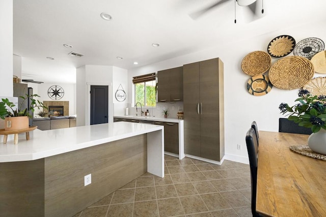 kitchen featuring ceiling fan, backsplash, dark brown cabinetry, a tile fireplace, and sink