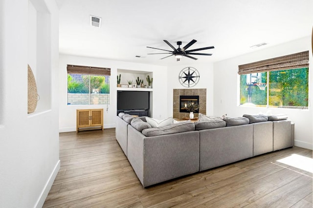 living room featuring ceiling fan, built in shelves, a tiled fireplace, and light hardwood / wood-style flooring