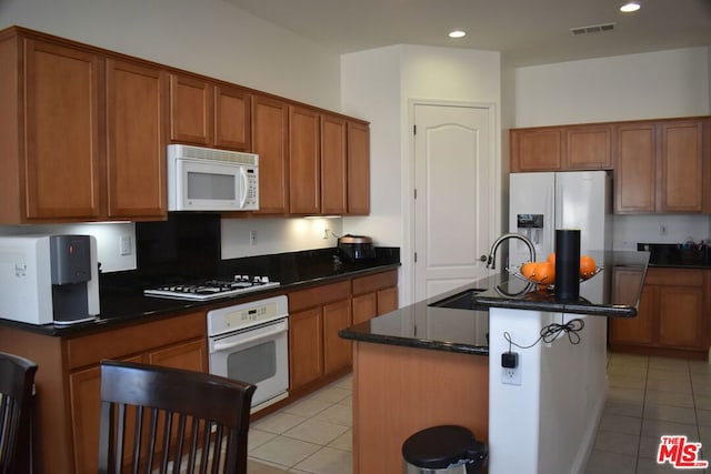 kitchen featuring a center island with sink, white appliances, light tile patterned flooring, dark stone counters, and sink