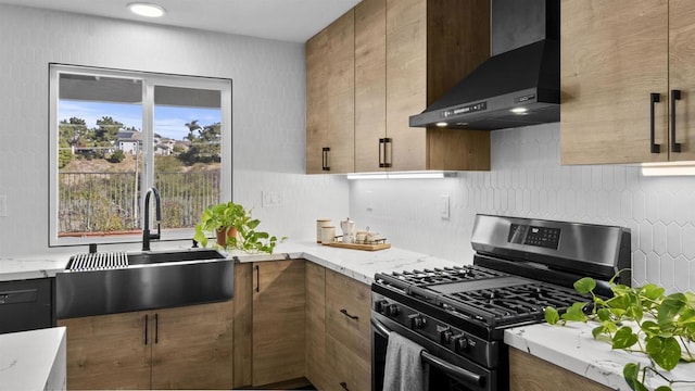 kitchen featuring stainless steel gas range oven, tasteful backsplash, wall chimney range hood, light stone counters, and sink