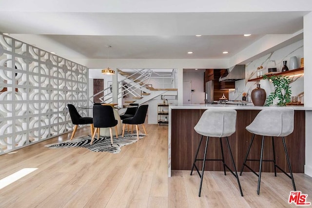 kitchen with wall chimney range hood, a kitchen bar, kitchen peninsula, light hardwood / wood-style flooring, and hanging light fixtures