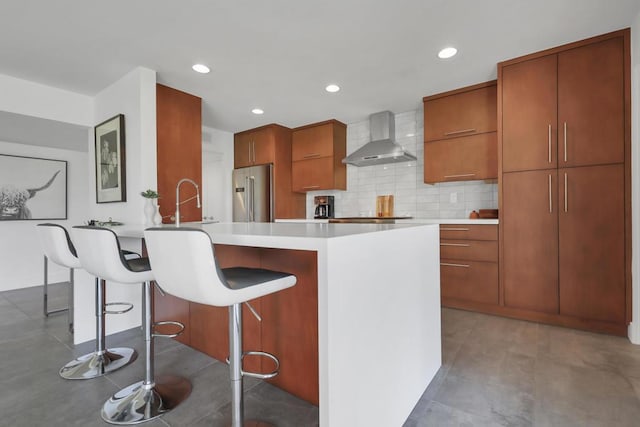 kitchen featuring high end fridge, backsplash, a kitchen island with sink, a kitchen breakfast bar, and wall chimney exhaust hood