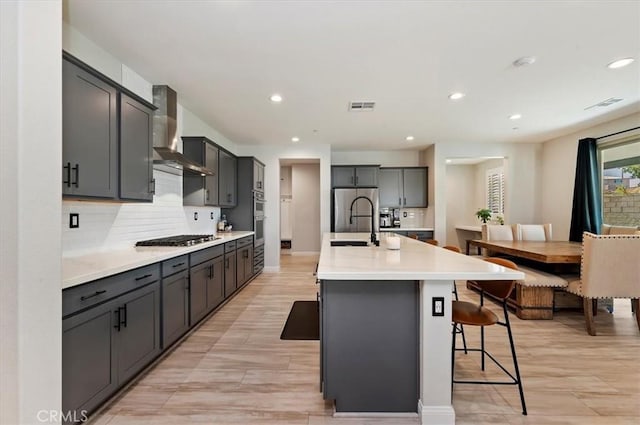 kitchen with gray cabinets, an island with sink, appliances with stainless steel finishes, tasteful backsplash, and wall chimney range hood