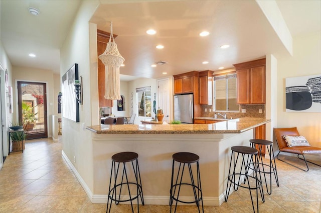 kitchen featuring decorative backsplash, kitchen peninsula, stainless steel refrigerator, light stone counters, and a breakfast bar