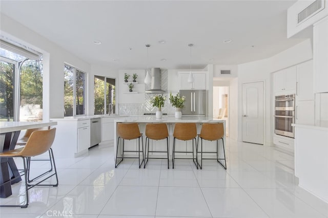 kitchen with wall chimney range hood, appliances with stainless steel finishes, white cabinetry, a center island, and decorative light fixtures