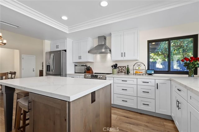 kitchen featuring light stone countertops, white cabinets, appliances with stainless steel finishes, wall chimney exhaust hood, and crown molding