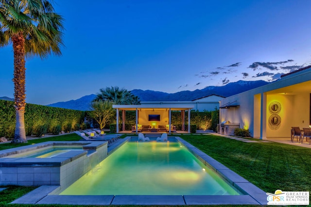 pool at dusk with an in ground hot tub, a lawn, a mountain view, and an outdoor living space with a fireplace