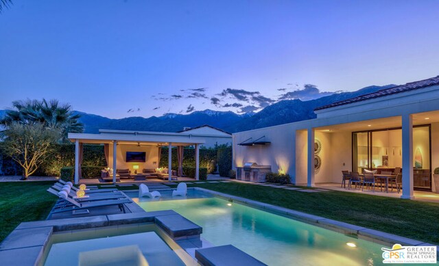 pool at dusk with a mountain view, a lawn, an outdoor living space with a fireplace, an in ground hot tub, and a patio