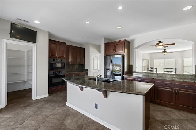 kitchen featuring stainless steel fridge with ice dispenser, multiple ovens, a center island with sink, ceiling fan, and a kitchen breakfast bar