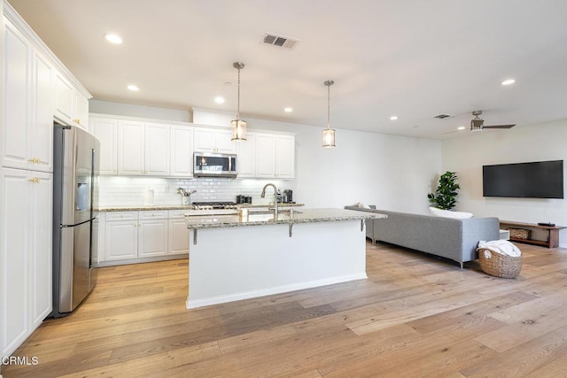 kitchen featuring pendant lighting, white cabinets, appliances with stainless steel finishes, an island with sink, and light stone counters