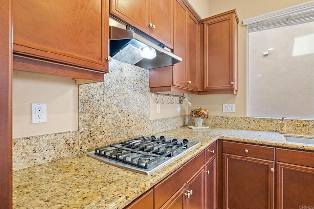 kitchen featuring stainless steel gas cooktop, light stone counters, brown cabinets, and under cabinet range hood