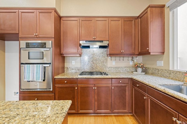 kitchen with light stone counters, light wood-style flooring, under cabinet range hood, stainless steel appliances, and brown cabinets