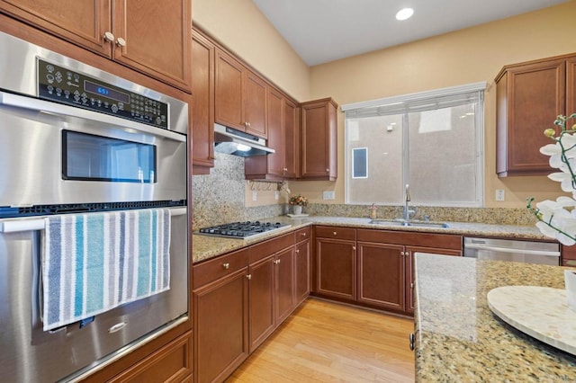 kitchen with light stone counters, brown cabinets, appliances with stainless steel finishes, a sink, and under cabinet range hood