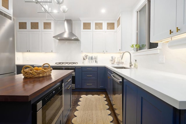 kitchen with wall chimney range hood, sink, white cabinets, blue cabinets, and wood counters