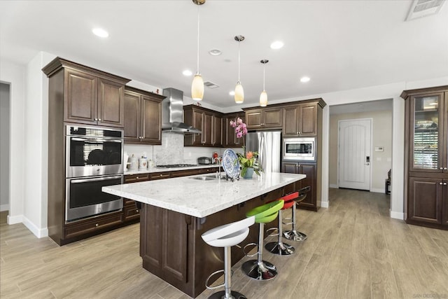 kitchen with wall chimney range hood, a center island with sink, dark brown cabinetry, hanging light fixtures, and appliances with stainless steel finishes