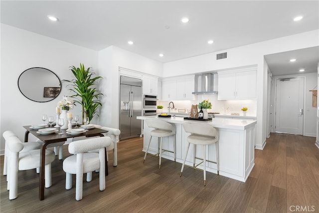 kitchen with appliances with stainless steel finishes, wall chimney exhaust hood, white cabinets, and a kitchen island with sink