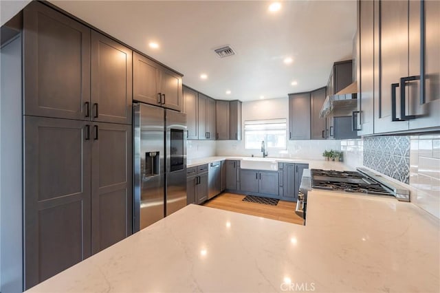 kitchen featuring stainless steel appliances, tasteful backsplash, wall chimney exhaust hood, light stone counters, and sink