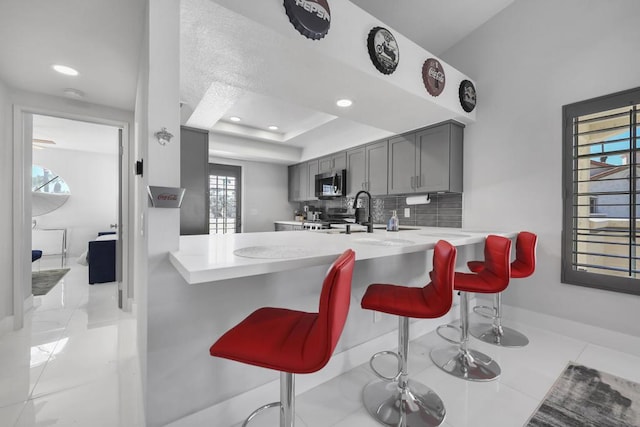 kitchen with a raised ceiling, kitchen peninsula, a kitchen breakfast bar, gray cabinetry, and stainless steel appliances