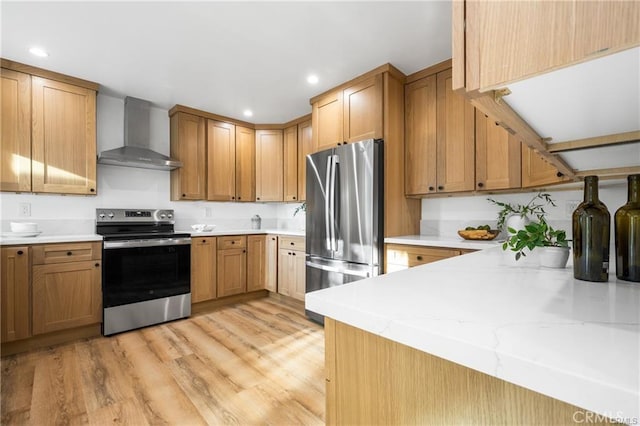 kitchen featuring wall chimney range hood, light hardwood / wood-style floors, kitchen peninsula, light stone countertops, and appliances with stainless steel finishes