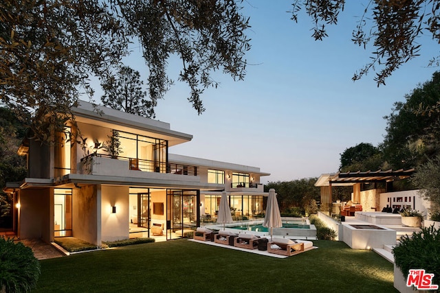 back house at dusk featuring a balcony, outdoor lounge area, a patio, a pool with hot tub, and a yard