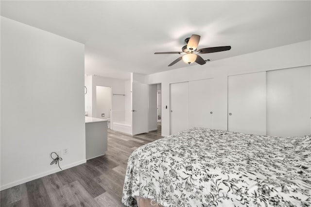 bedroom featuring two closets, a ceiling fan, ensuite bath, wood finished floors, and baseboards