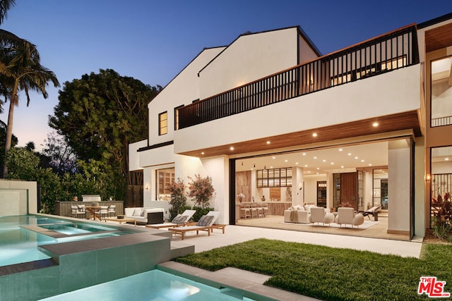 back house at dusk with a pool with hot tub, a patio area, exterior kitchen, a balcony, and an outdoor hangout area