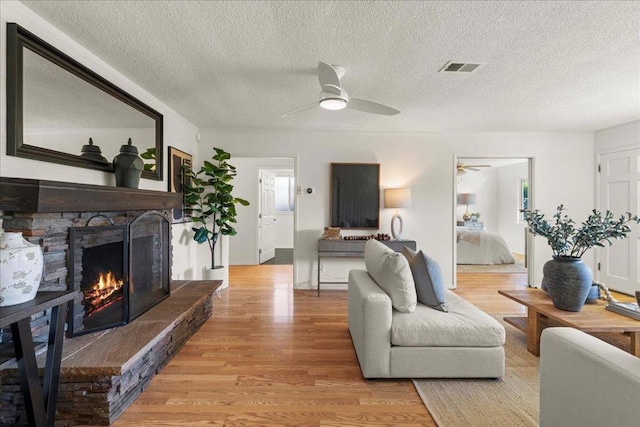 living room with a stone fireplace, a textured ceiling, ceiling fan, and light wood-type flooring