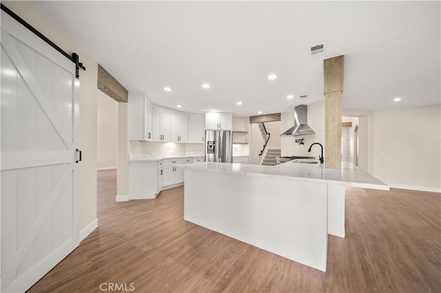 kitchen featuring a barn door, wall chimney range hood, white cabinetry, light hardwood / wood-style flooring, and stainless steel fridge with ice dispenser