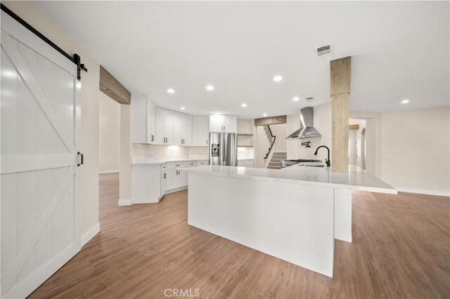 kitchen featuring wall chimney range hood, stainless steel refrigerator with ice dispenser, a barn door, light wood finished floors, and white cabinets
