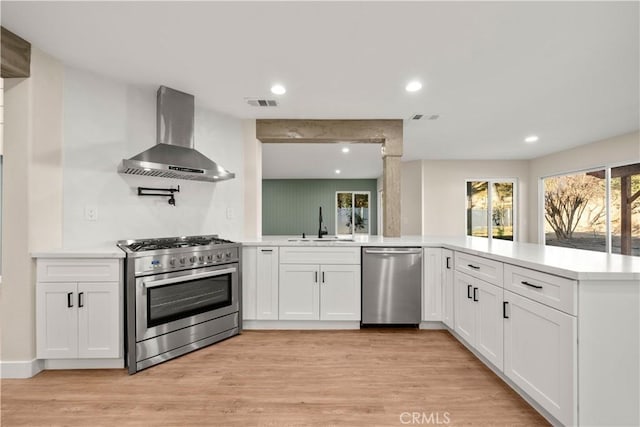 kitchen with kitchen peninsula, wall chimney range hood, white cabinets, and stainless steel appliances