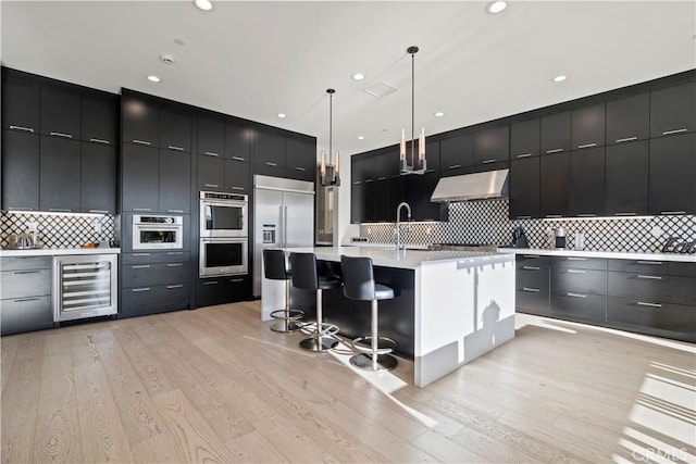 kitchen featuring appliances with stainless steel finishes, decorative light fixtures, wall chimney range hood, wine cooler, and a kitchen island with sink