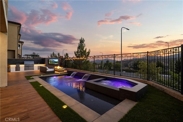 pool at dusk featuring pool water feature, an outdoor kitchen, area for grilling, and an in ground hot tub