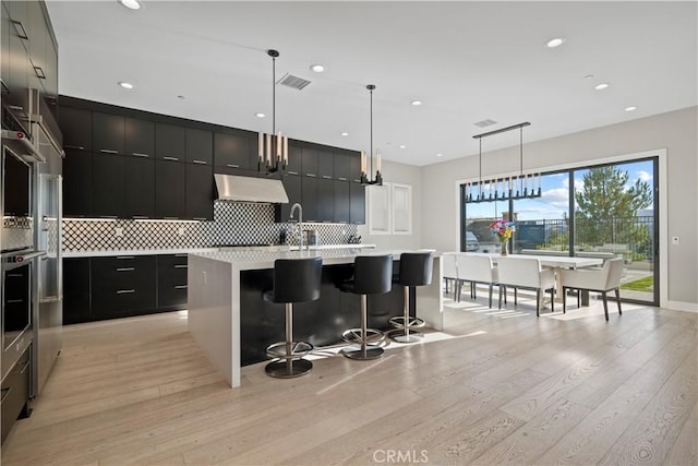 kitchen featuring an island with sink, light hardwood / wood-style floors, decorative backsplash, hanging light fixtures, and a breakfast bar area