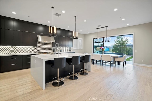 kitchen with extractor fan, a kitchen bar, hanging light fixtures, a kitchen island with sink, and light wood-type flooring