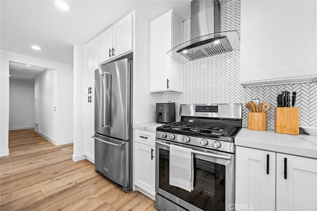 kitchen with appliances with stainless steel finishes, white cabinetry, wall chimney range hood, tasteful backsplash, and light hardwood / wood-style flooring