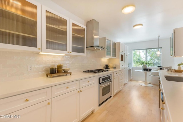 kitchen with appliances with stainless steel finishes, light hardwood / wood-style floors, white cabinets, decorative light fixtures, and wall chimney exhaust hood