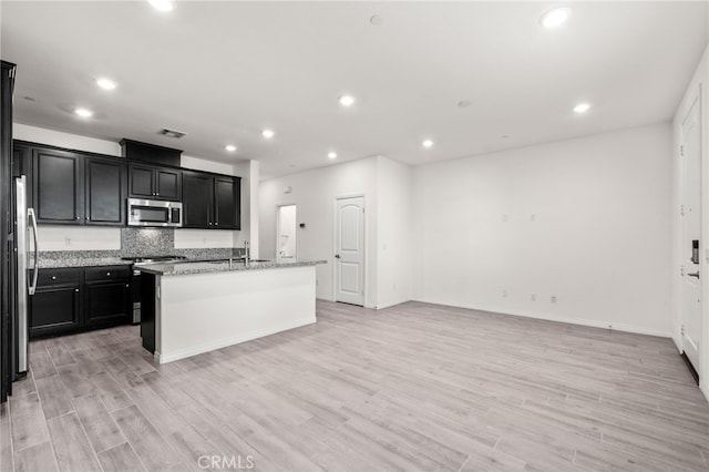 kitchen with light stone counters, light hardwood / wood-style flooring, stainless steel appliances, and an island with sink