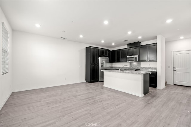 kitchen with light hardwood / wood-style floors, decorative backsplash, light stone countertops, an island with sink, and stainless steel appliances