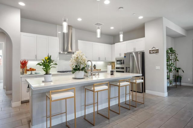 kitchen with white cabinets, appliances with stainless steel finishes, wall chimney exhaust hood, an island with sink, and hanging light fixtures