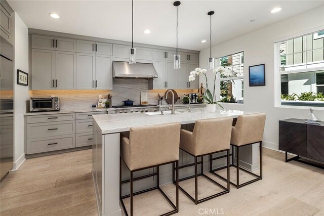 kitchen with a center island with sink, ventilation hood, gray cabinetry, decorative light fixtures, and light stone counters