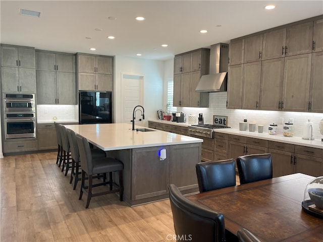 kitchen featuring a kitchen bar, sink, appliances with stainless steel finishes, an island with sink, and wall chimney exhaust hood