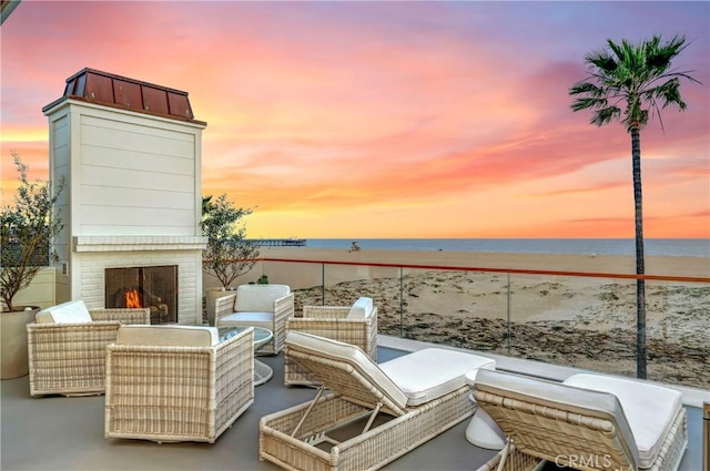 patio terrace at dusk with a water view, an outdoor brick fireplace, and a view of the beach