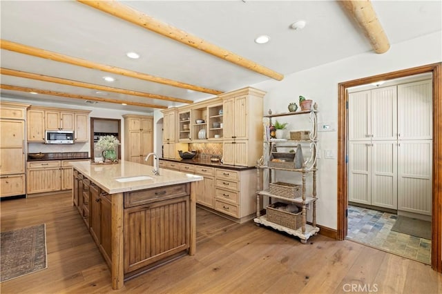 kitchen featuring wood-type flooring, an island with sink, sink, light stone counters, and beam ceiling