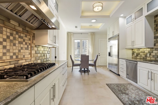 kitchen featuring white cabinetry, stainless steel appliances, a raised ceiling, wall chimney range hood, and stone counters