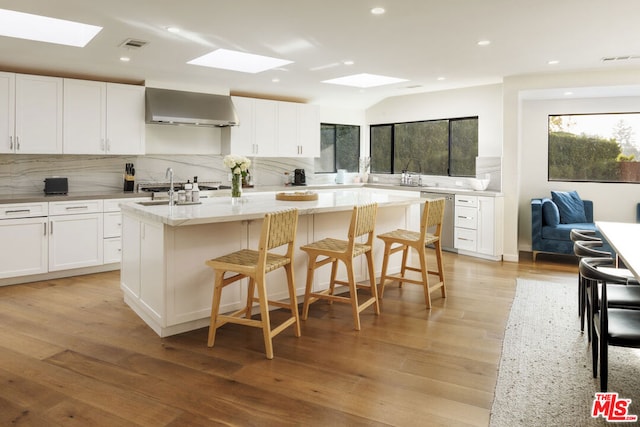 kitchen with a skylight, white cabinets, a center island with sink, and wall chimney exhaust hood