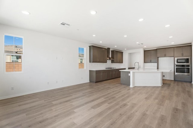 kitchen featuring wall chimney exhaust hood, an island with sink, sink, oven, and light wood-type flooring